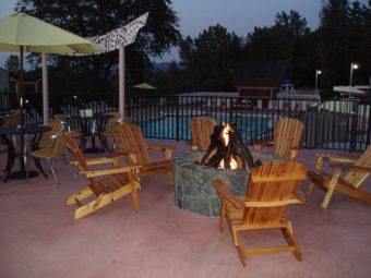 Lit fire pit with wood and flames, wooden pool chairs, parasols and distant view of outdoor pool behind fencing
